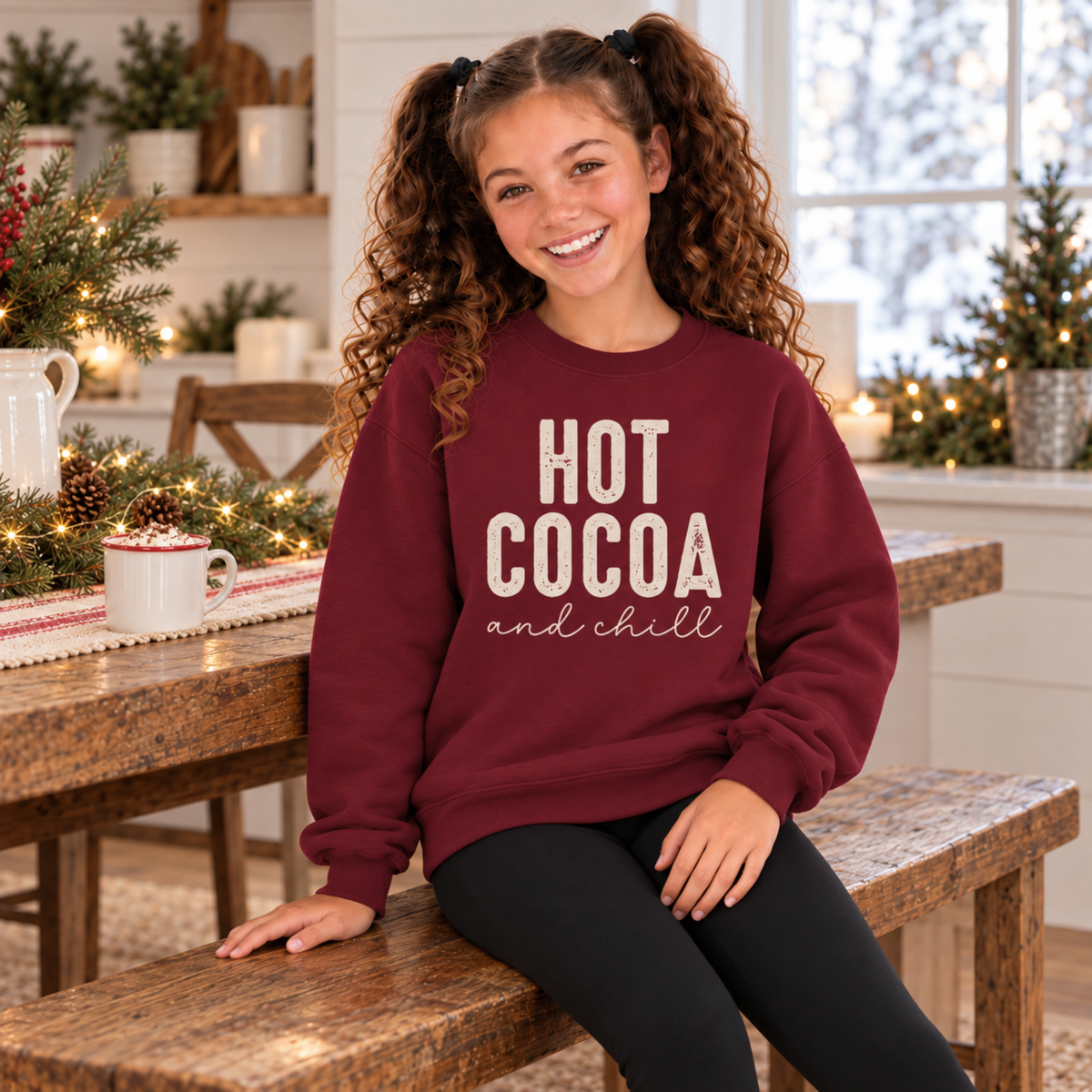 Girl wearing a maroon youth sweatshirt with Hot Cocoa and Chill design in a festive winter kitchen