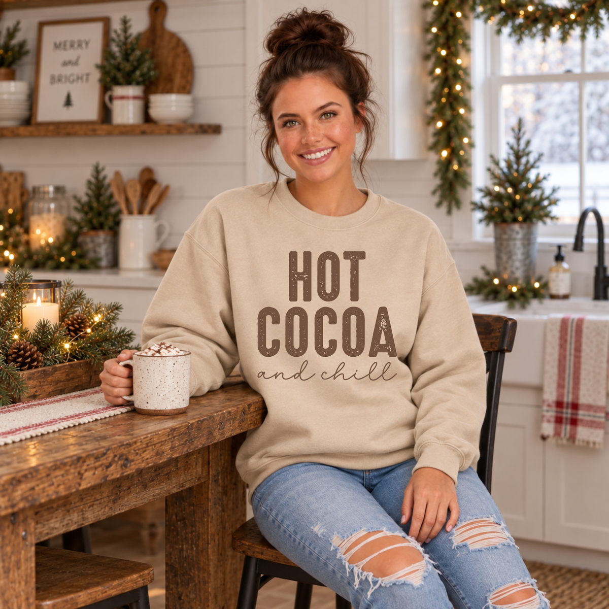 Woman in a sand Hot Cocoa and Chill sweatshirt holding a cocoa mug at a rustic farmhouse table