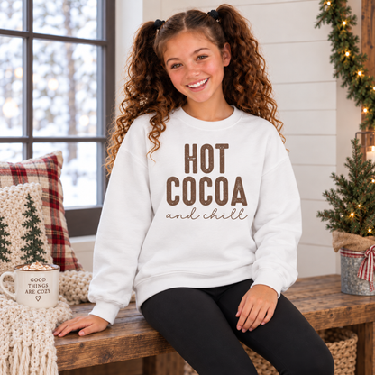 Girl wearing a white youth sweatshirt with brown Hot Cocoa and Chill design in a cozy winter room