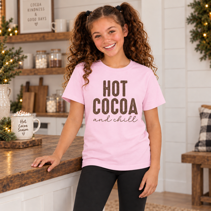 Girl wearing a light pink youth T-shirt with brown Hot Cocoa and Chill design beside a mug of cocoa