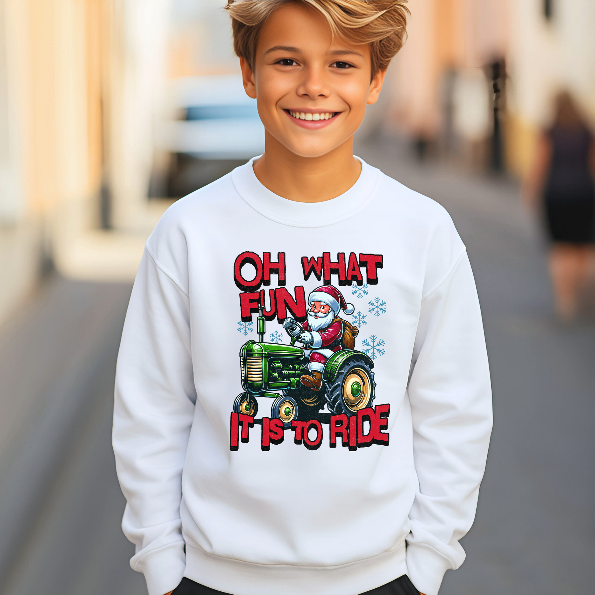 Smiling boy wearing a white youth Christmas sweatshirt with Santa riding a green tractor and the words “Oh What Fun It Is to Ride.”