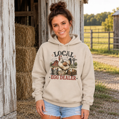 Woman wearing a sand Local Egg Dealer hoodie in front of a barn with hay bales.