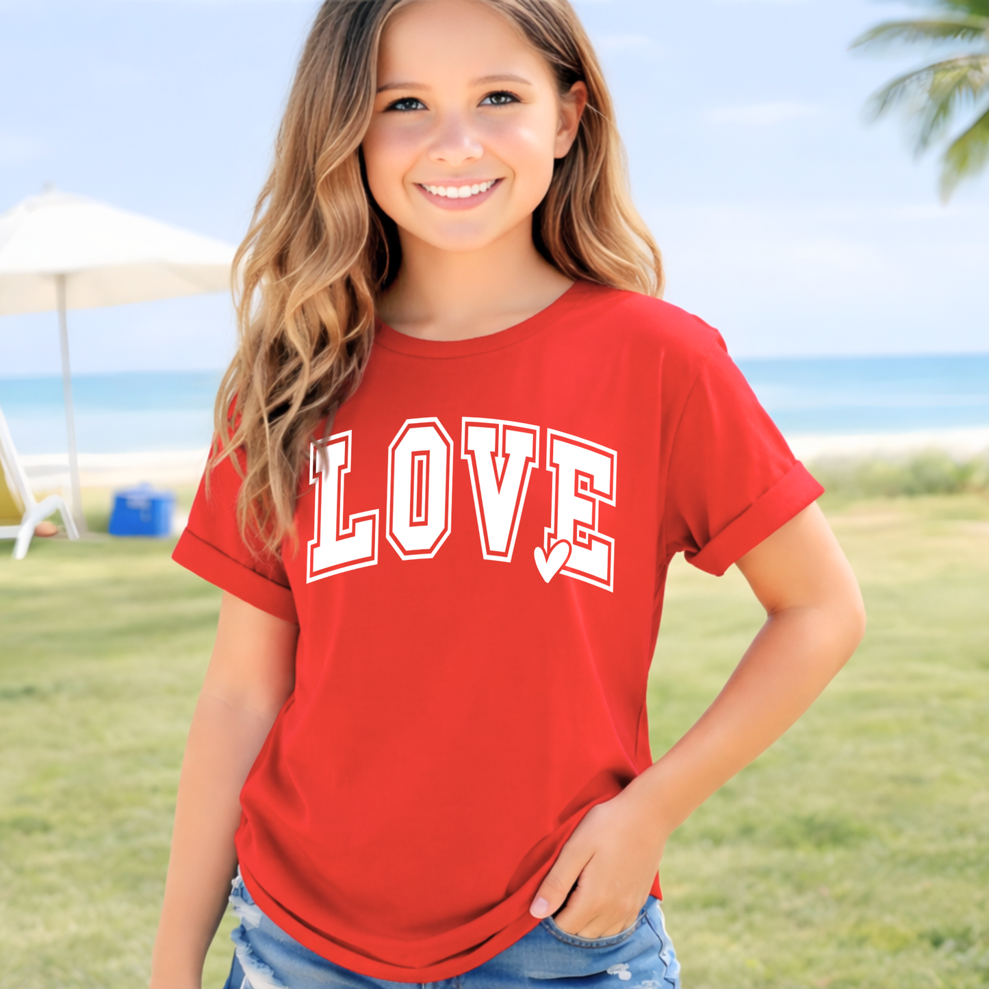 Youth girls red LOVE graphic t-shirt with white varsity lettering and heart detail, styled on beach with denim shorts, summer kids outfit, Valentine shirt, casual youth apparel