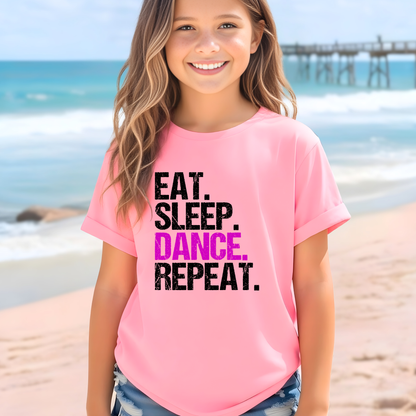 Young girl wearing a pink t-shirt with 'EAT. SLEEP. DANCE. REPEAT.' text on a beach.