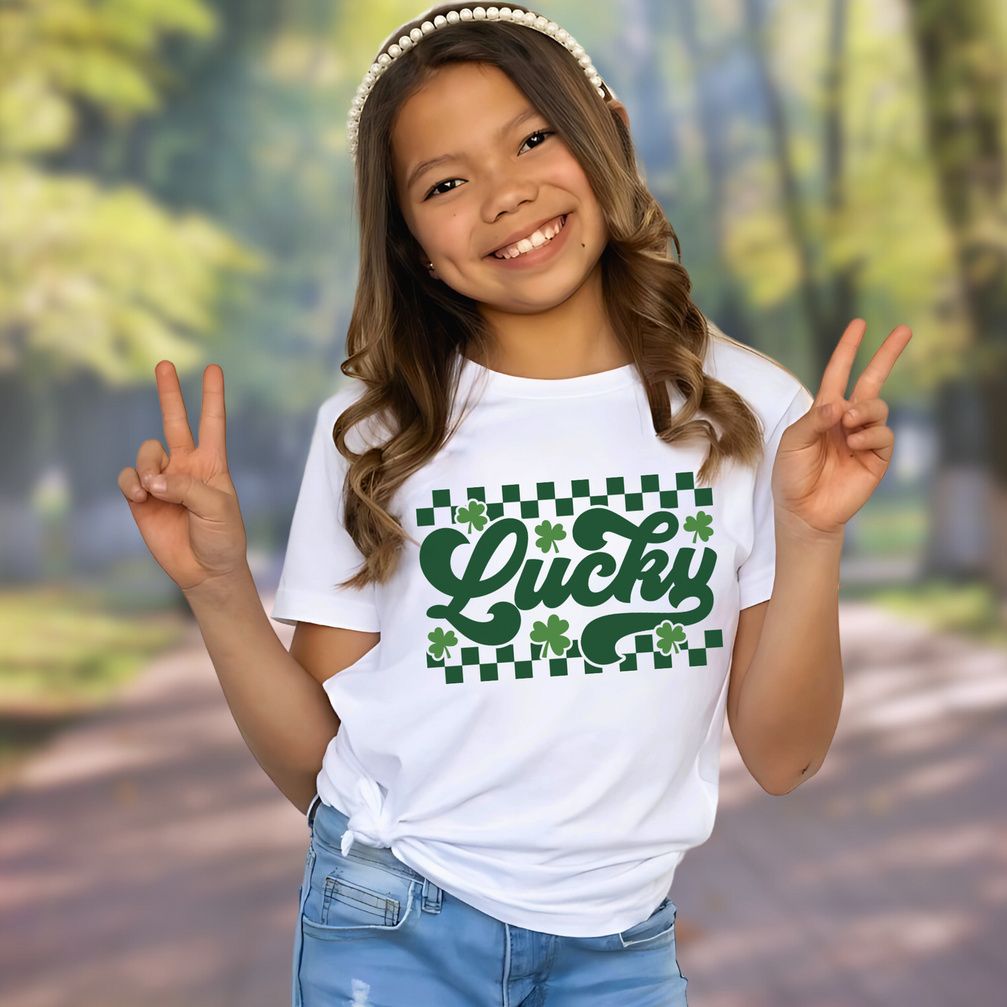 White T-shirt with 'Lucky' in green cursive, surrounded by shamrocks and a checkered pattern – perfect for St. Patrick’s Day.
