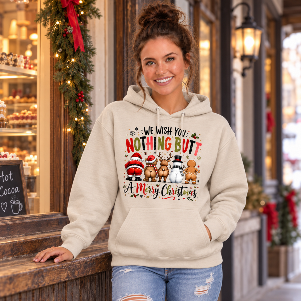 Woman wearing a sand hoodie with We Wish You Nothing Butt a Merry Christmas design with rear-view Santa, bear, reindeer, snowman and gingerbread characters outside a decorated holiday bakery