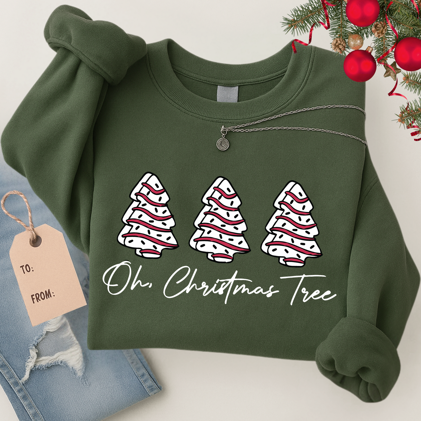 Flat lay of a green Christmas sweatshirt with three festive trees and “Oh Christmas Tree” script. Styled with jeans, necklace, and holiday gift tag for a cozy seasonal look.