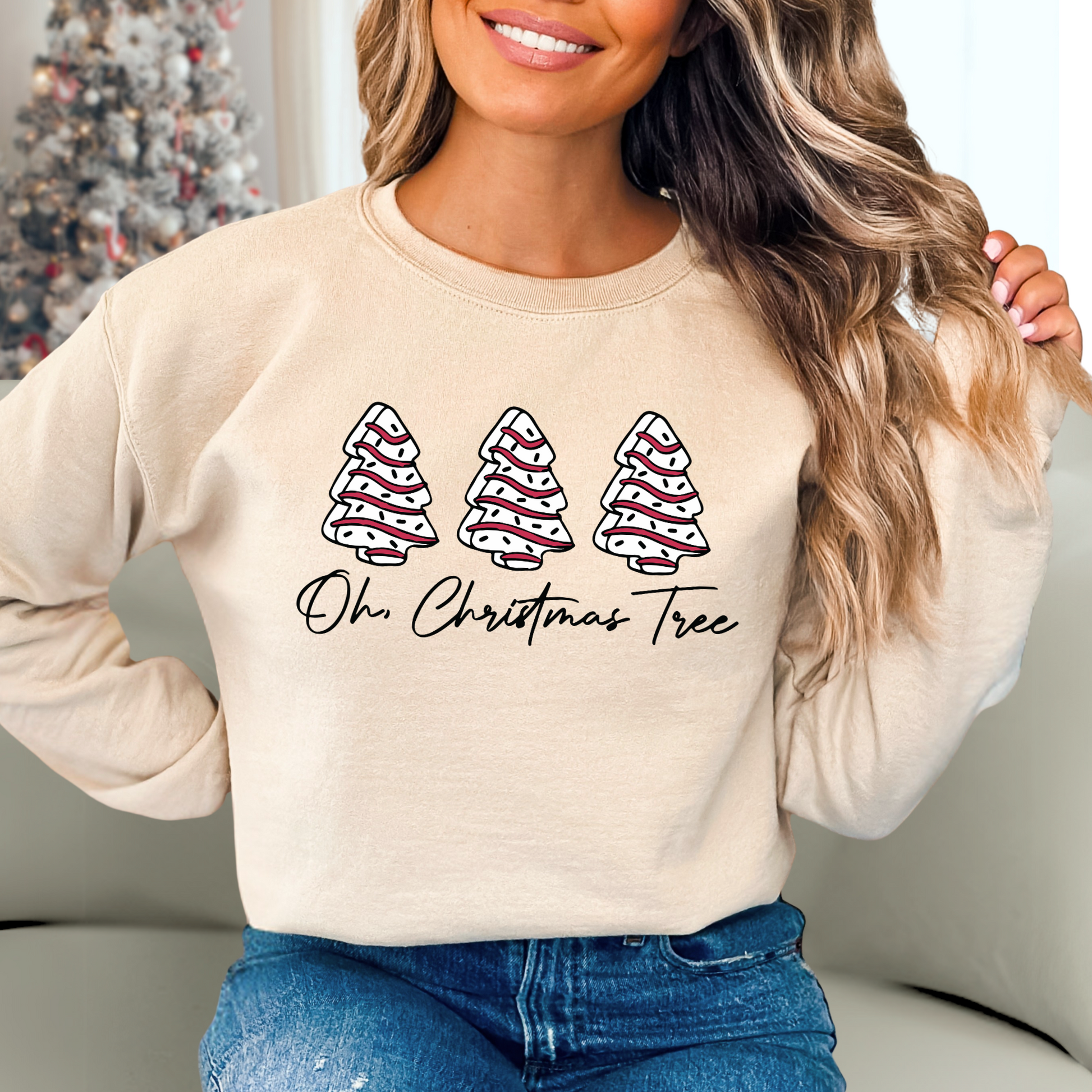 Woman wearing a sand color unisex Christmas sweatshirt with three festive white and red trees and the phrase “Oh Christmas Tree.” Cozy holiday pullover styled with jeans.