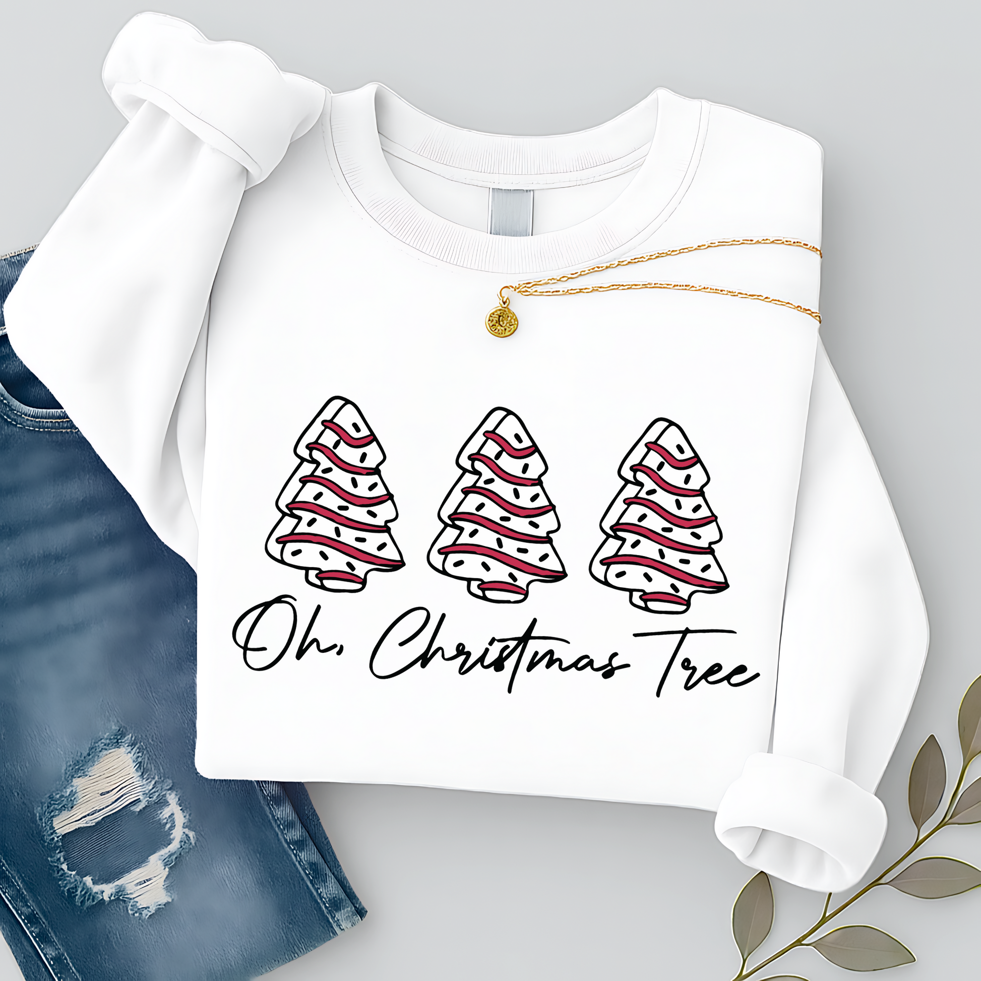Flat lay of a white Christmas sweatshirt featuring three red and white trees with “Oh Christmas Tree” text. Styled with ripped jeans and gold necklace for a festive look.