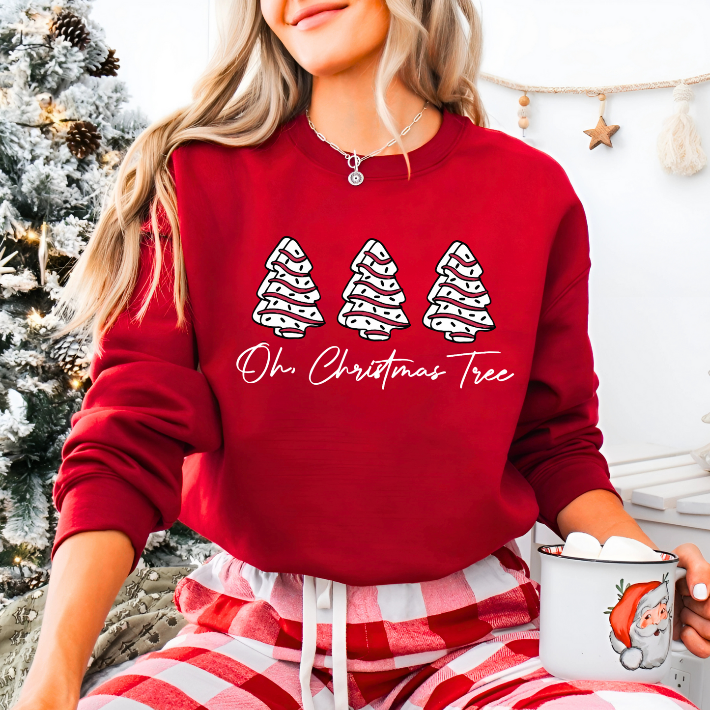 Woman wearing a red unisex Christmas sweatshirt with three decorated trees and “Oh Christmas Tree” text. Cozy holiday outfit styled with plaid pants and a Santa mug.