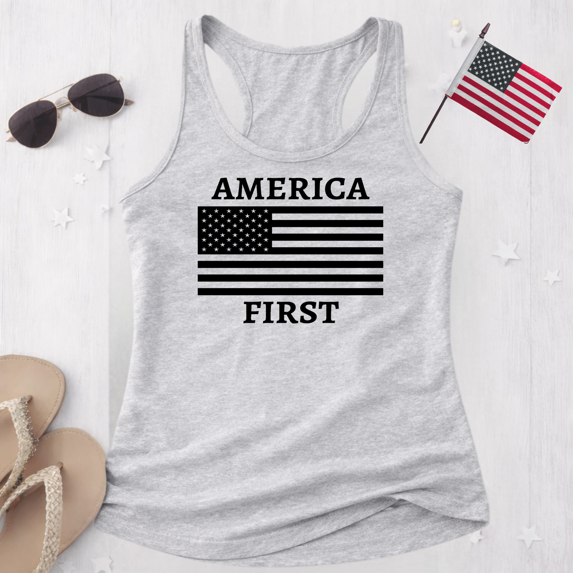 Gray tank top with 'AMERICA FIRST' text and American flag design on a white surface with sunglasses and an American flag.