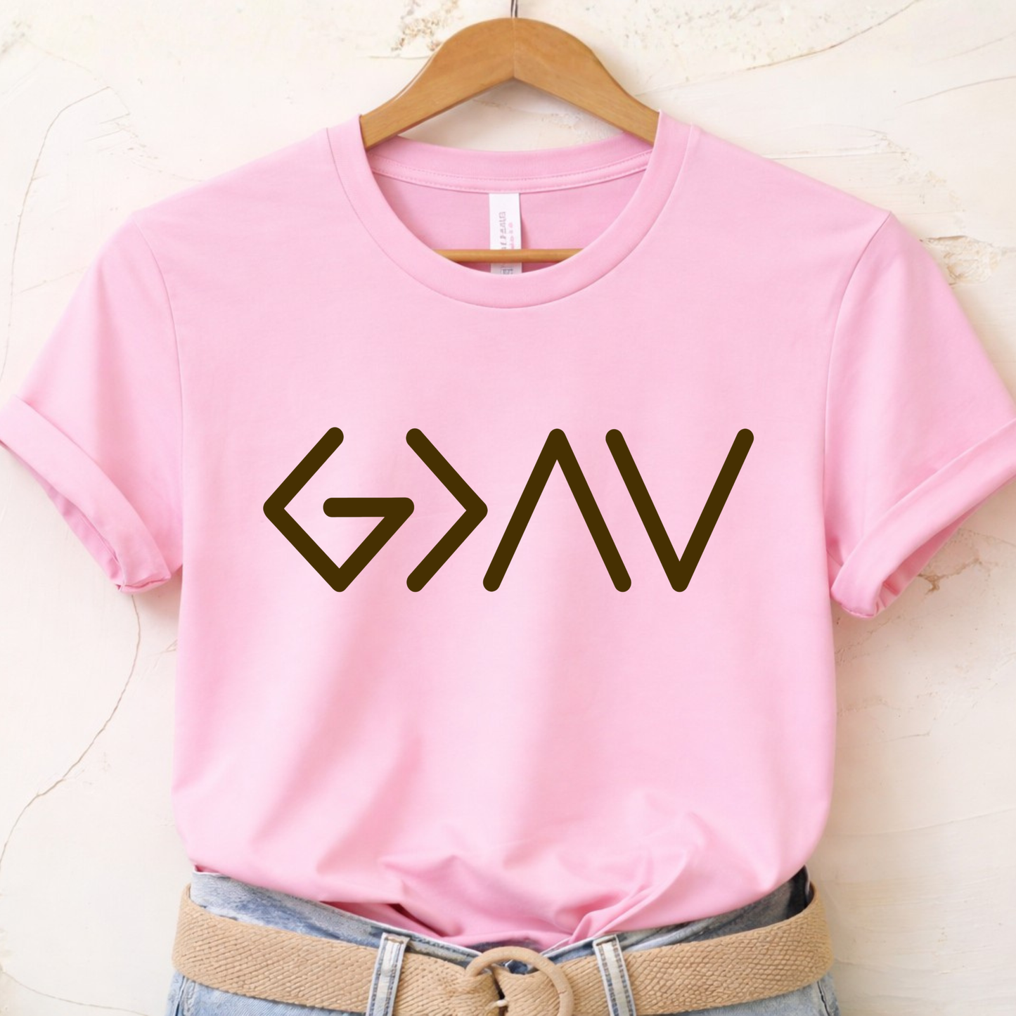 Pink short sleeve t-shirt folded featuring gold God Is Greater Than the Highs and Lows front design
