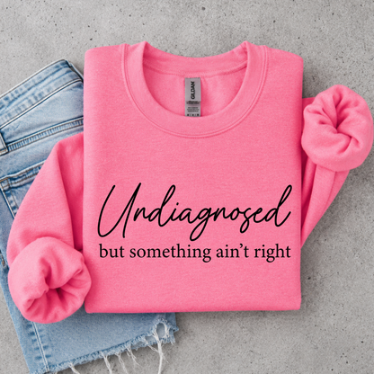 Bright pink crewneck sweatshirt flat lay with black Undiagnosed but something ain’t right graphic text.