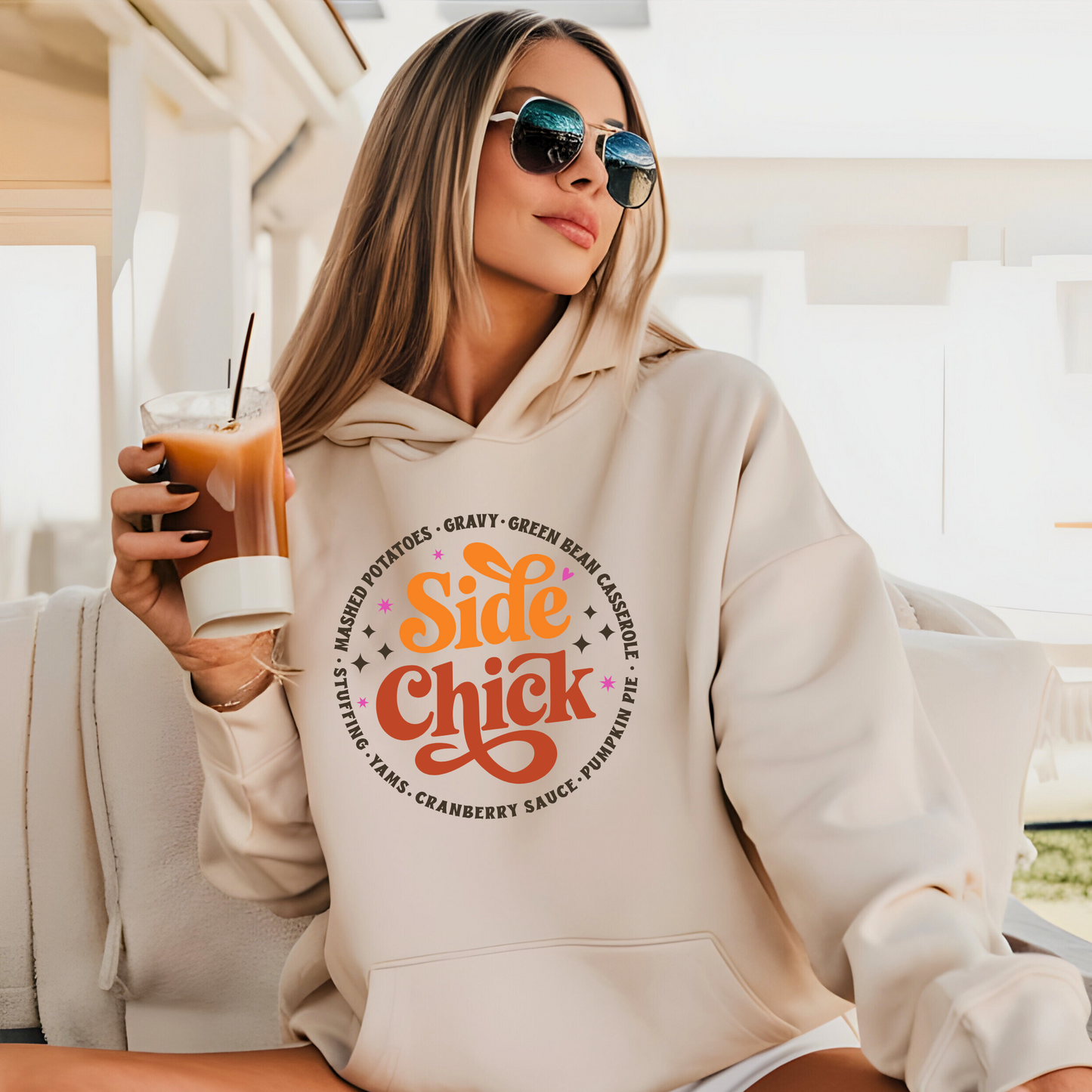 Trendy woman in light sand “Side Chick” hoodie holding iced coffee outdoors. Cozy Thanksgiving hoodie styled for casual fall looks and perfect for holiday travel or lounging.