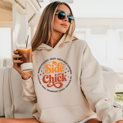 Trendy woman in light sand “Side Chick” hoodie holding iced coffee outdoors. Cozy Thanksgiving hoodie styled for casual fall looks and perfect for holiday travel or lounging.