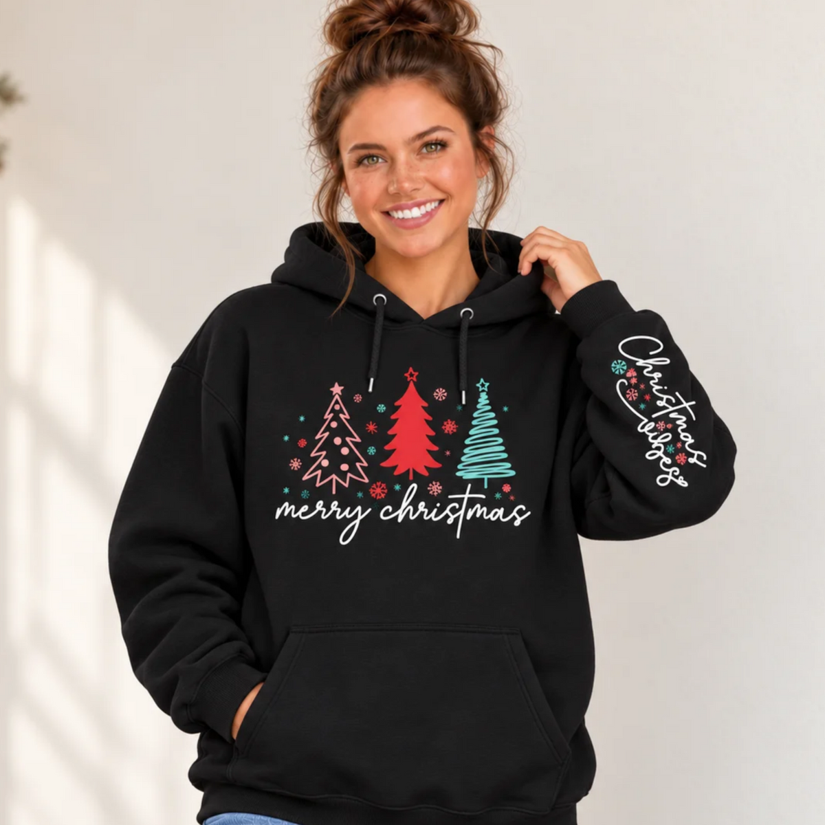 Woman wearing a black hoodie with pink, red, and teal Christmas trees, Merry Christmas front design, and Christmas Vibes sleeve print