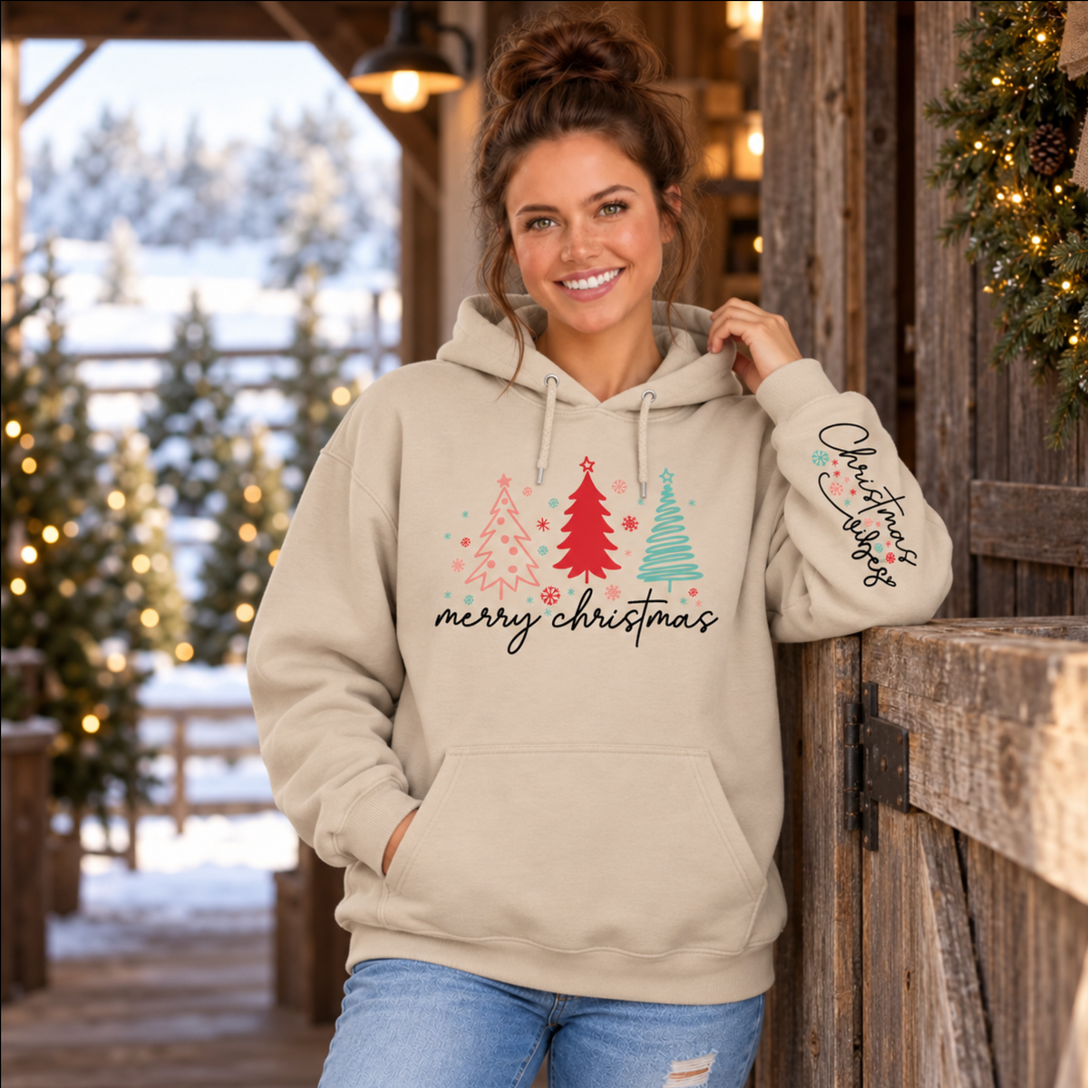Woman wearing a sand Merry Christmas tree hoodie with Christmas Vibes sleeve print beside a rustic barn and snowy trees