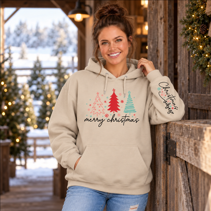 Woman wearing a sand Merry Christmas tree hoodie with Christmas Vibes sleeve print beside a rustic barn and snowy trees