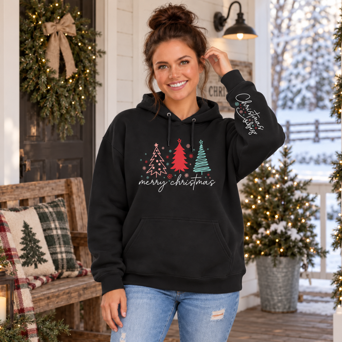 Woman wearing a black Merry Christmas tree hoodie with Christmas Vibes sleeve print on a snowy decorated porch