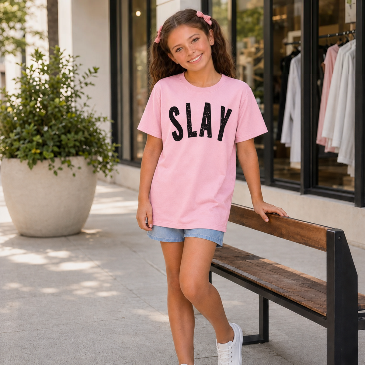 Young girl wearing a light pink short-sleeve SLAY T-shirt with bold black distressed lettering beside an outdoor bench.