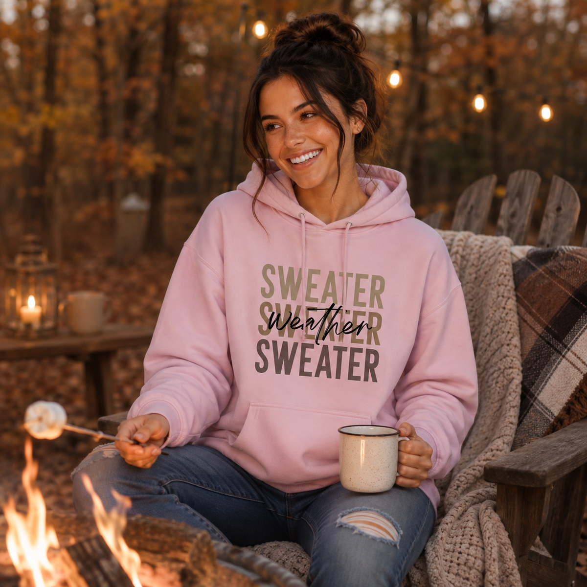 Woman wearing light pink Sweater Weather hoodie by a fall campfire