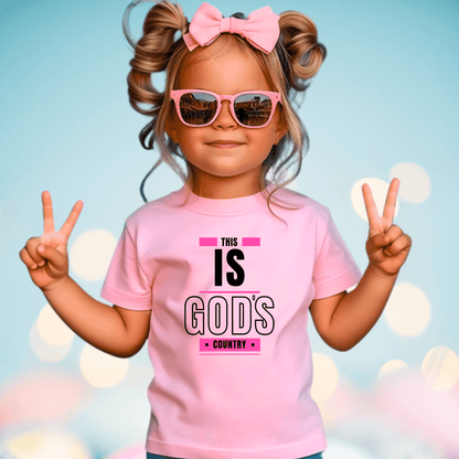 Smiling girl in a light pink 'This Is God's Country' youth t-shirt, a stylish and inspirational Christian kids' shirt.