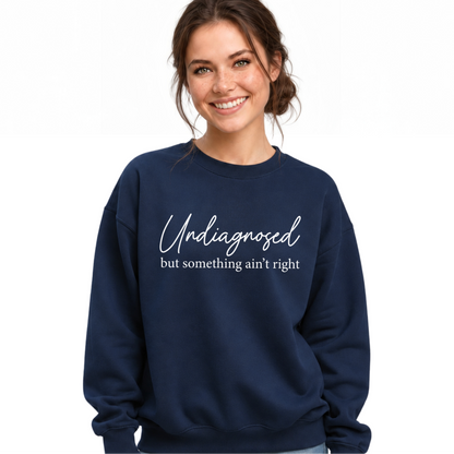 A woman wearing a navy blue crewneck sweatshirt with the white printed text 'Undiagnosed but something ain't right' on the chest, paired with distressed light-wash jeans, against a plain white background.