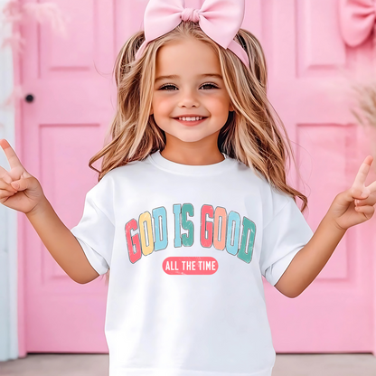Happy young girl wearing a white 'God Is Good – All the Time' Easter t-shirt, posing with peace signs in front of a pink Easter-themed background