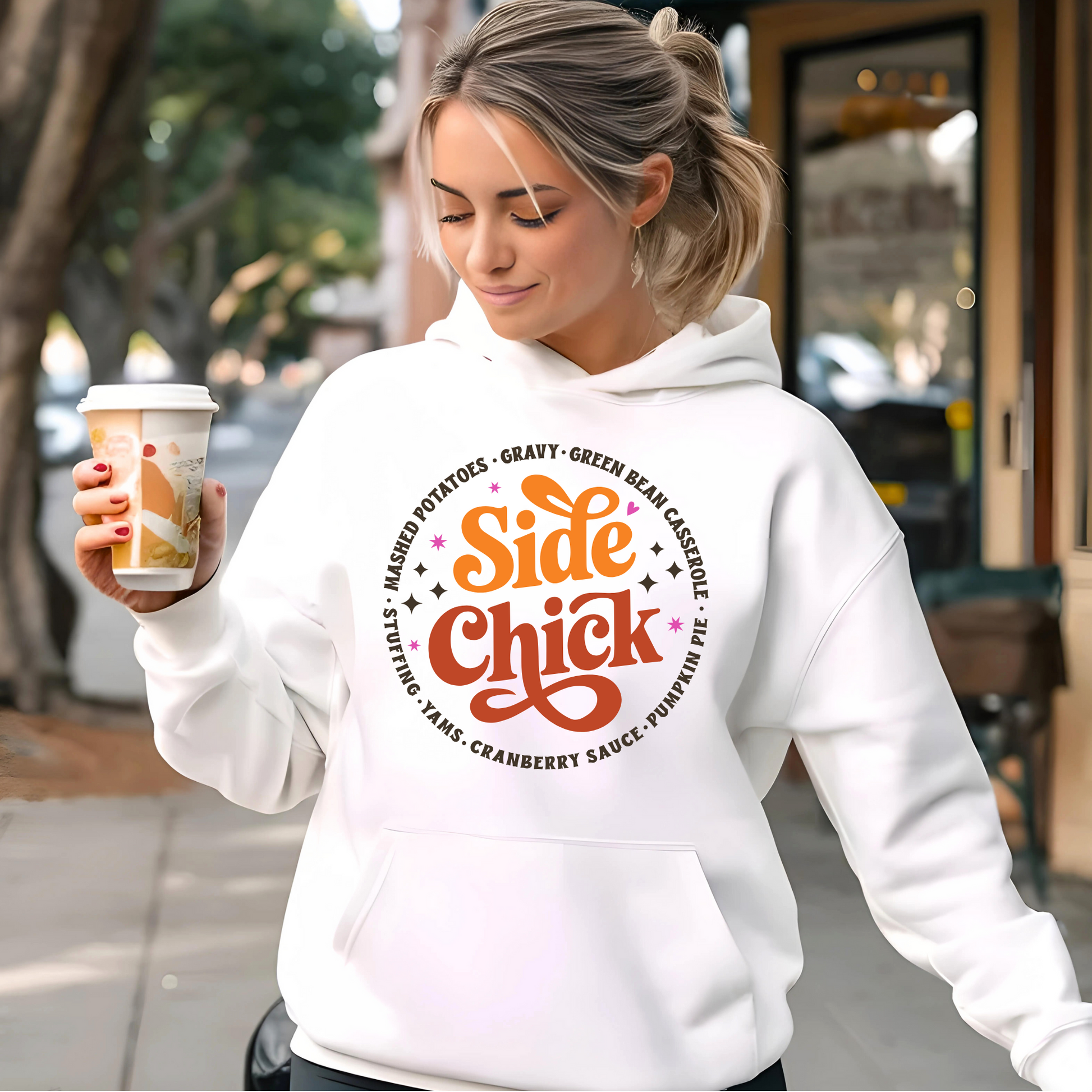 Woman holding coffee and wearing white “Side Chick” hoodie outside a cafe. Cute and cozy Thanksgiving hoodie with a food-themed design—perfect for holiday errands or pumpkin spice vibes.

